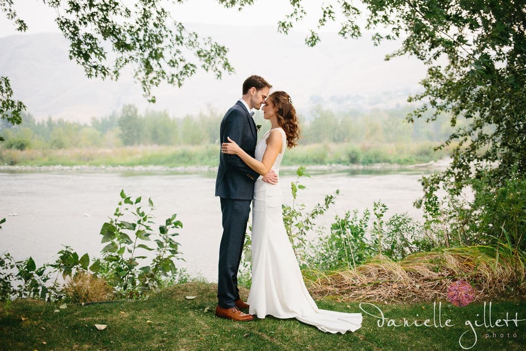A couple embraces by a serene river, surrounded by lush greenery.