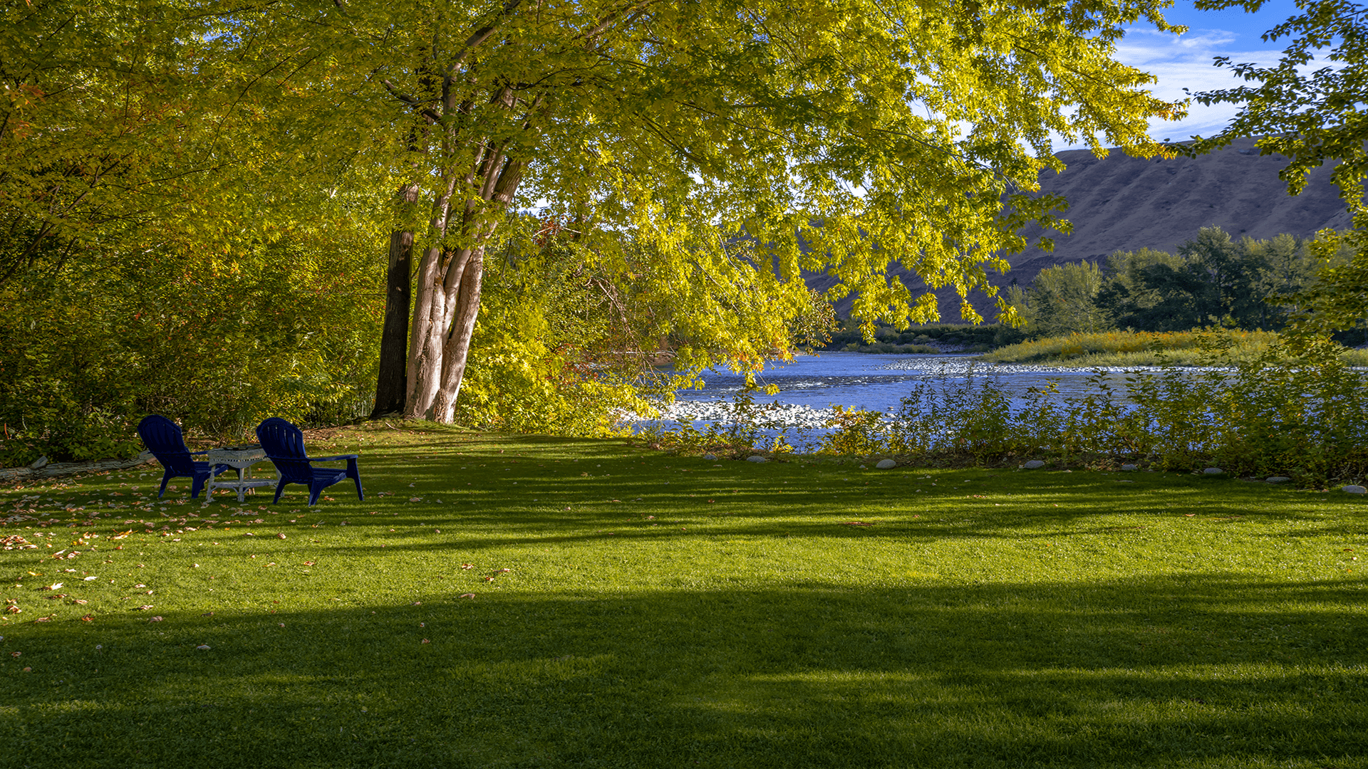Two blue chairs sit on a grassy bank beside a calm river, framed by lush trees and a distant hillside.
