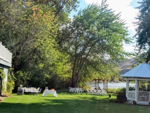 A picturesque outdoor venue featuring a green lawn, white chairs, a gazebo, and trees by a river.
