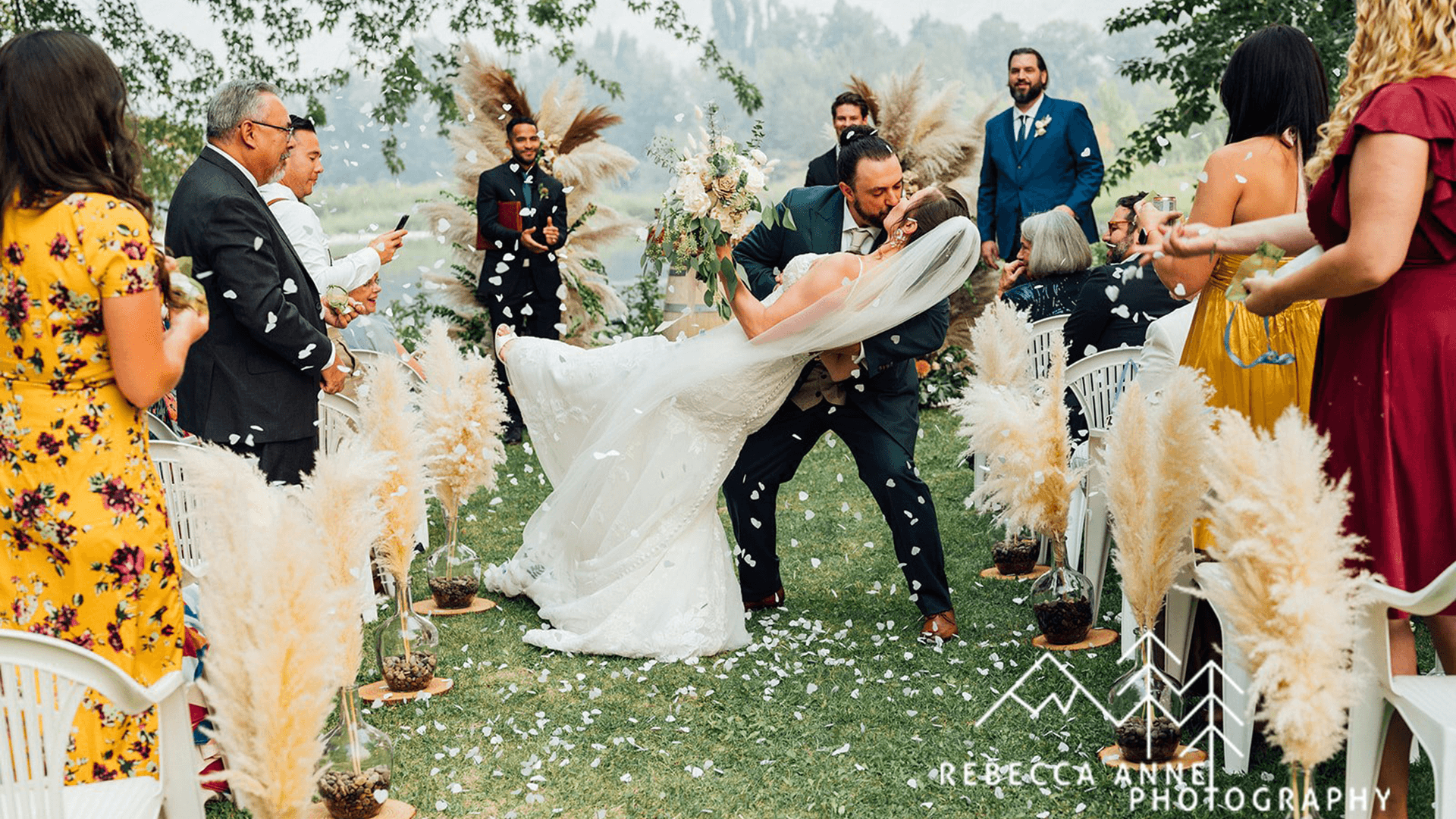 A couple shares a romantic kiss amid a wedding bouquet with guests celebrating around them.