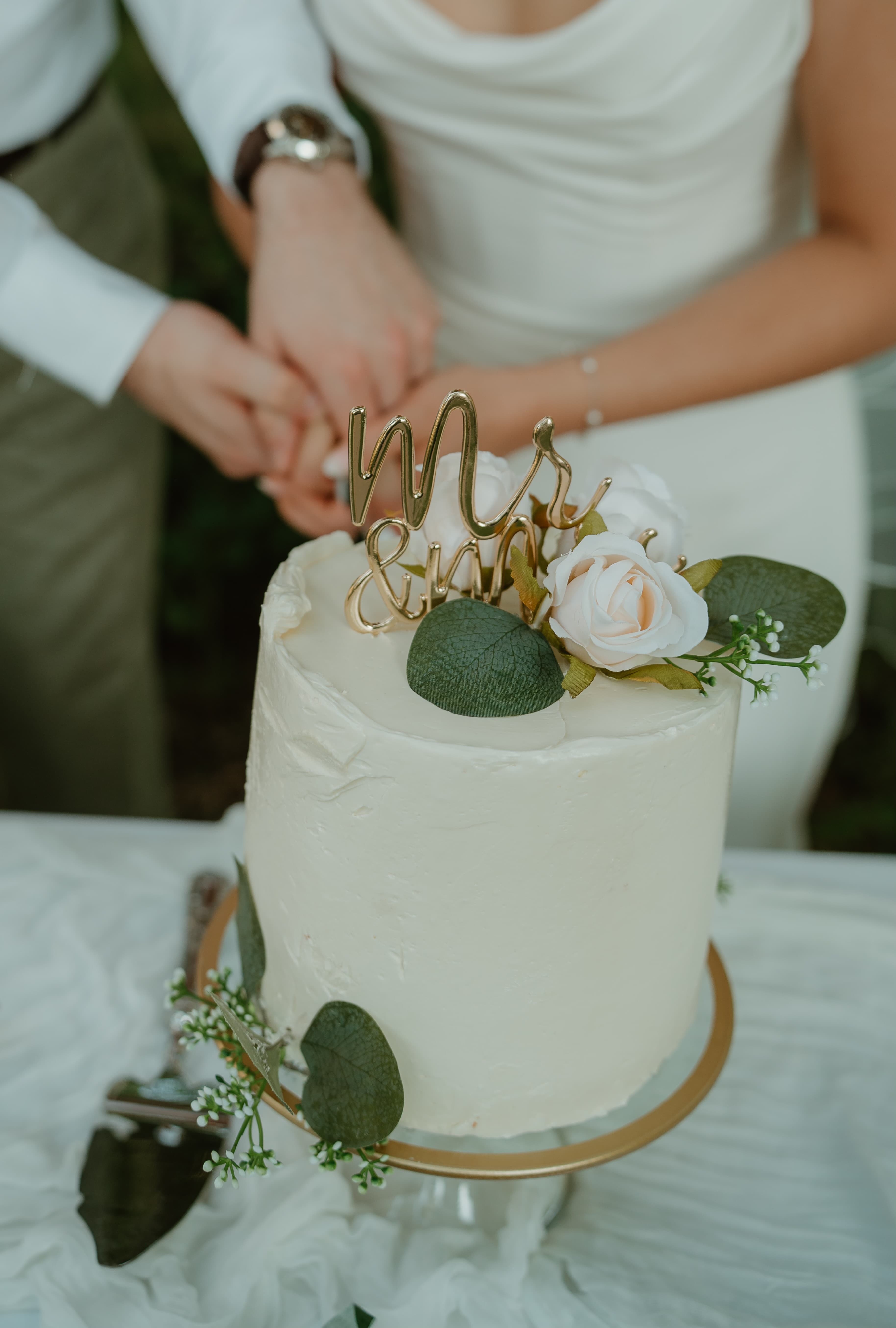 A wedding cake adorned with flowers and a "Mr & Mrs" topper, as a couple holds hands nearby.
