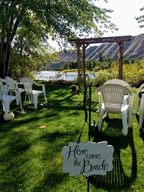 An outdoor wedding setup with chairs, a sign reading "Here comes the Bride," and a scenic backdrop of a river and hills.