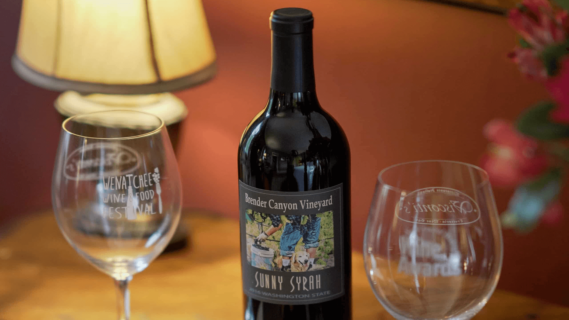 A bottle of Brender Canyon Vineyard Sunny Syrah wine is displayed alongside engraved wine glasses and a lamp.