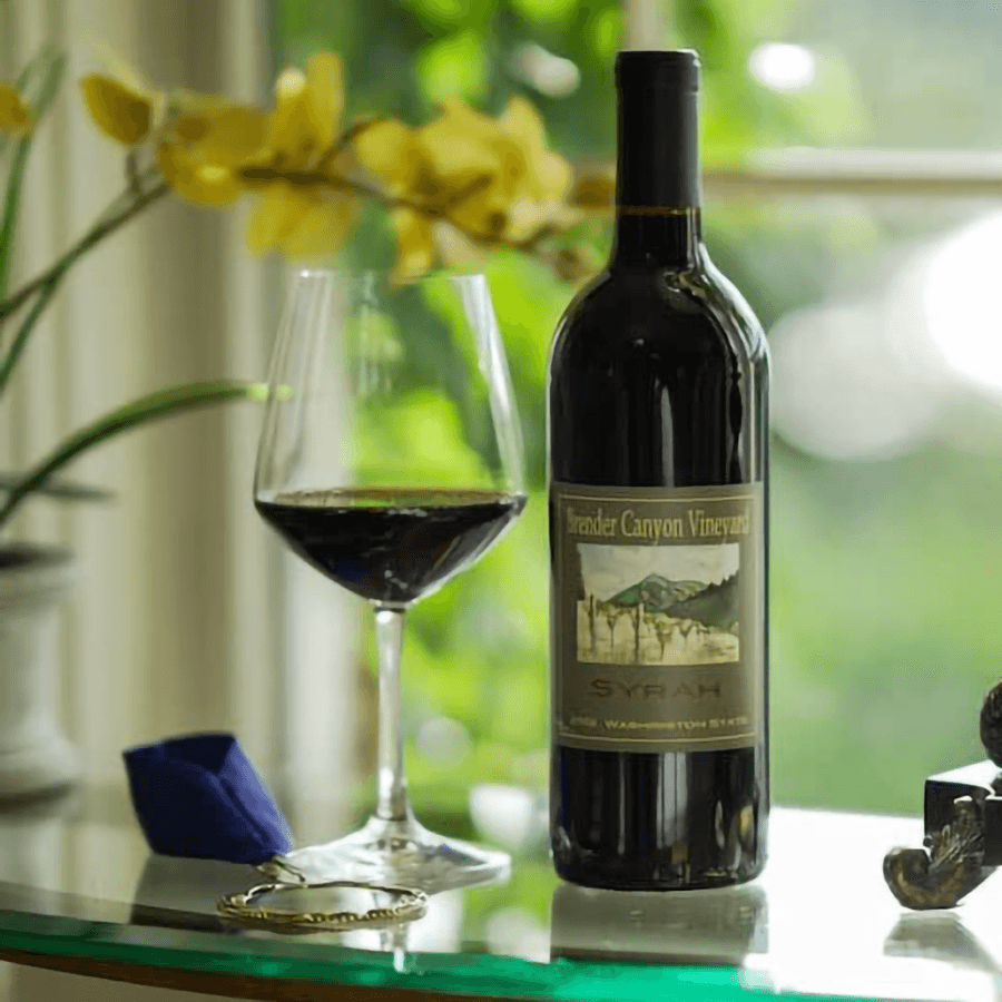 A bottle of Brander Canyon Vineyard Syrah next to a filled glass, with a blurred background of greenery and flowers.