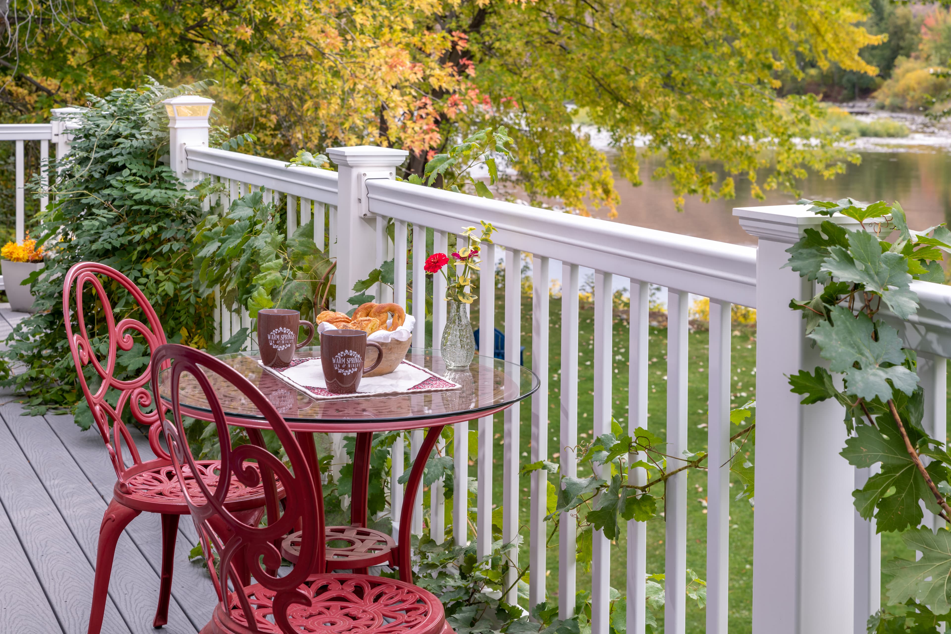 A cozy outdoor table with two red chairs, framed by greenery, features coffee mugs, pastries, and a flower vase overlooking a serene body of water.