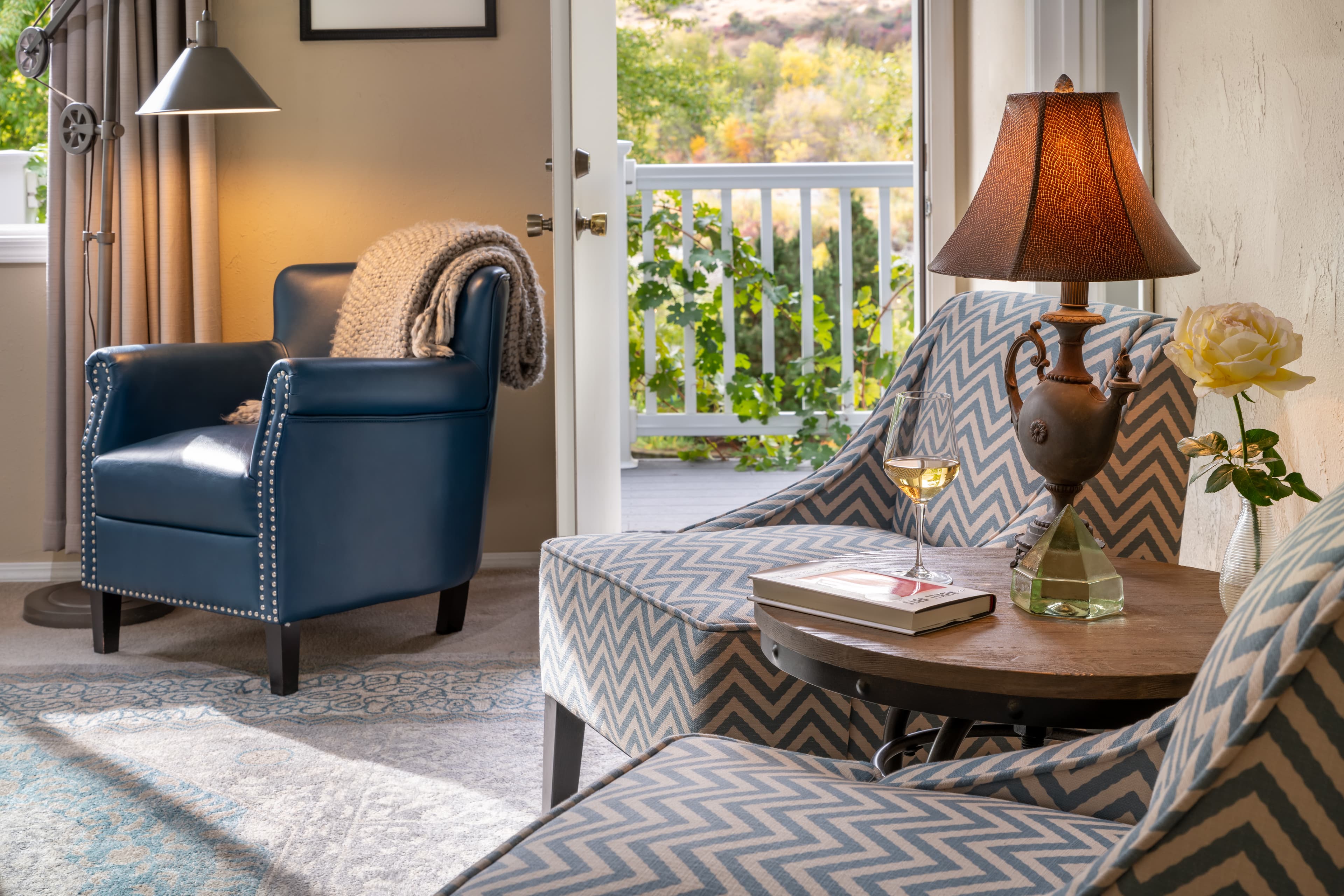 A cozy living space featuring patterned armchairs, a blue leather chair, a lamp, a small table with a drink, and a view of greenery outside.