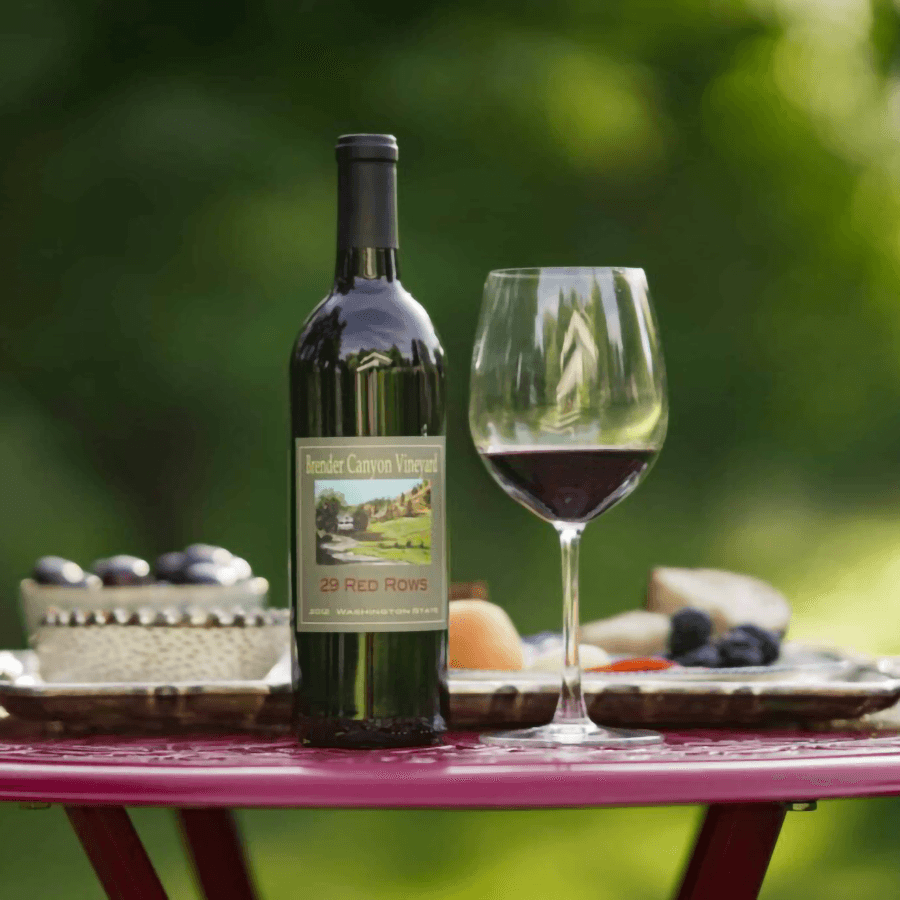A bottle of red wine with a glass, surrounded by fruits and a cake on a pink table.