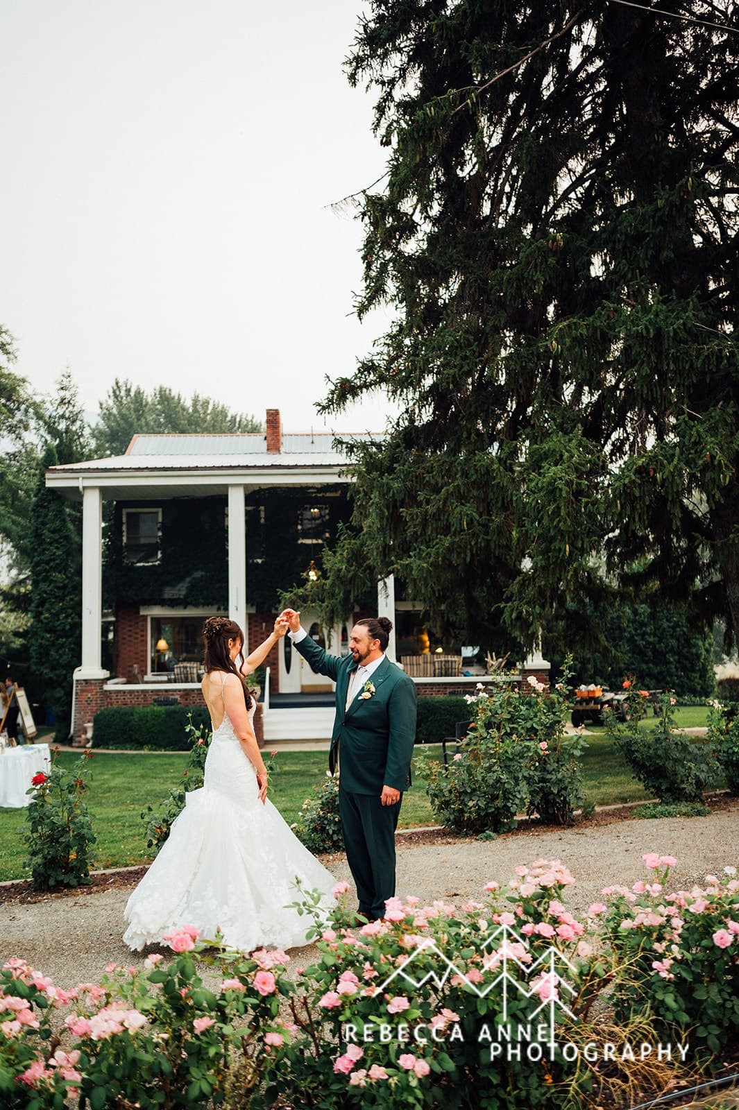 A bride in a white gown dances joyfully with a groom in a green suit in front of a quaint, ivy-covered house.