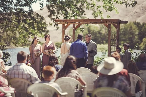 A couple exchanges vows during a wedding ceremony by a riverside, surrounded by guests.