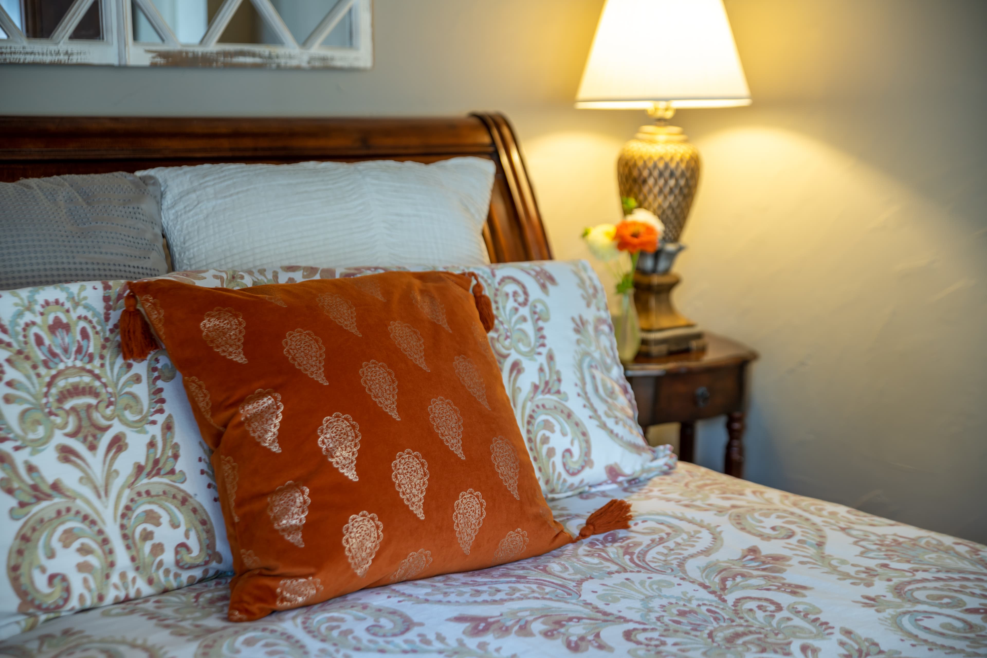 A cozy bedroom featuring an orange patterned pillow on a neatly made bed beside a lamp and decorative nightstand.
