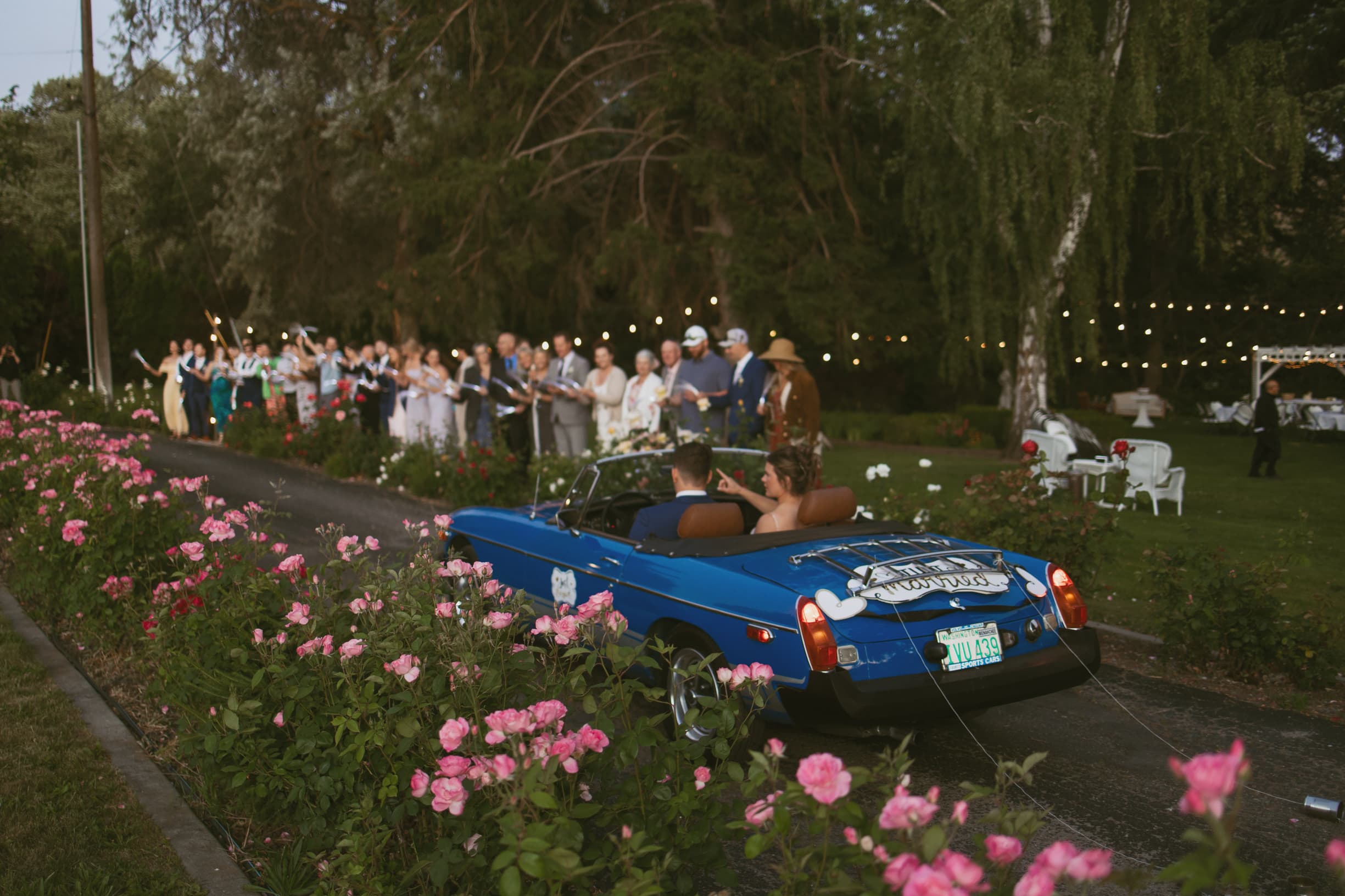 A couple drives away in a blue convertible surrounded by guests celebrating with sparklers in a garden.