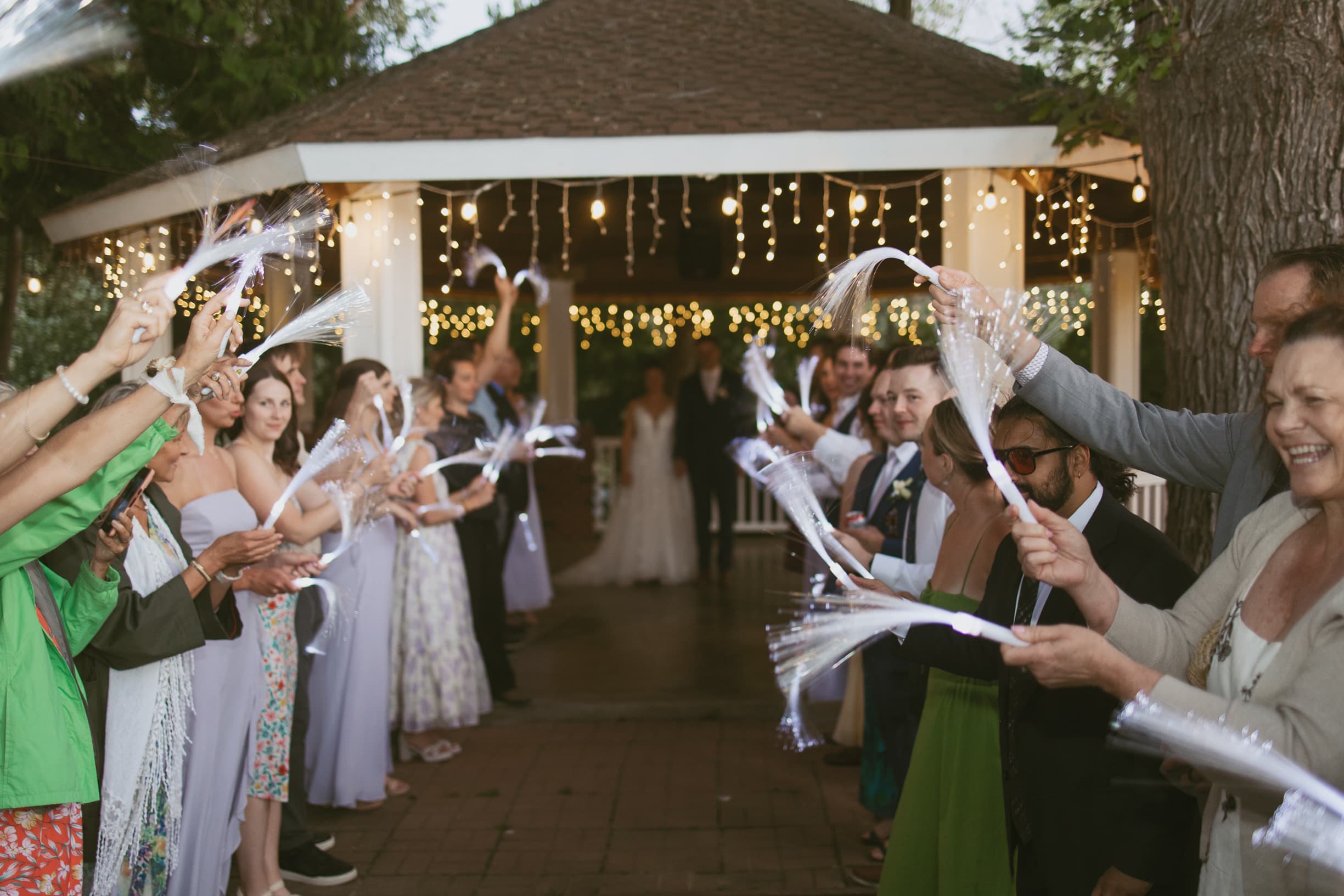 A group of wedding guests joyfully waves sparklers as the newlyweds exit a gazebo adorned with fairy lights.