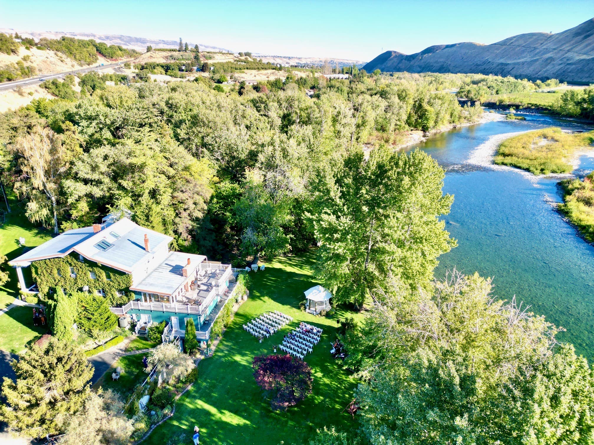 Aerial view of a scenic riverside venue with a house, lush greenery, and rows of white chairs set for an outdoor event.