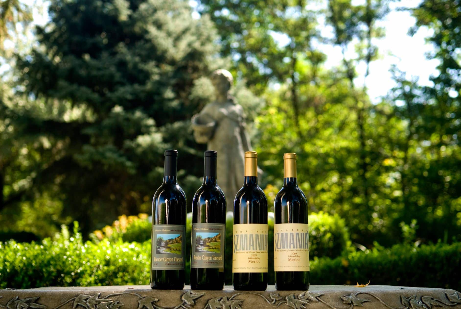 Four bottles of wine are displayed on a stone ledge, with a statue and lush greenery in the background.