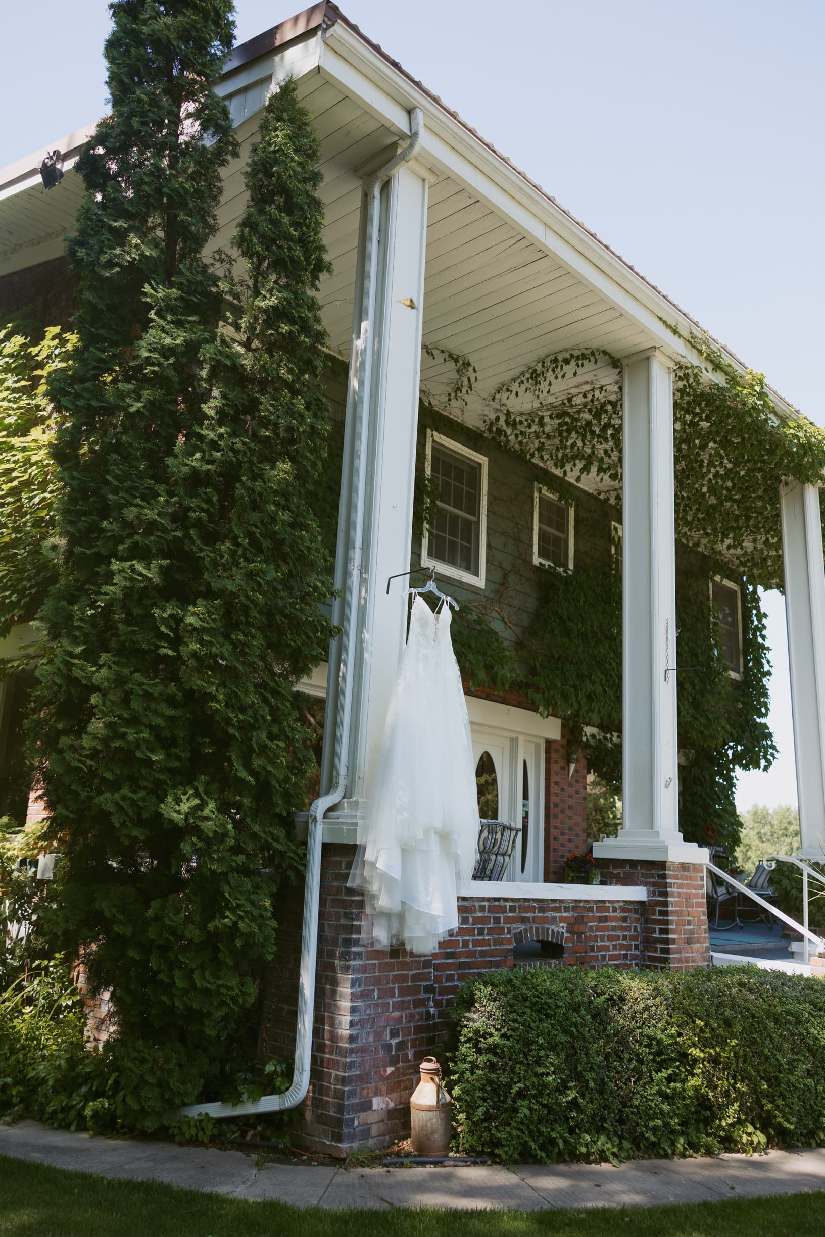 A white wedding dress hangs on a railing of a house surrounded by greenery.