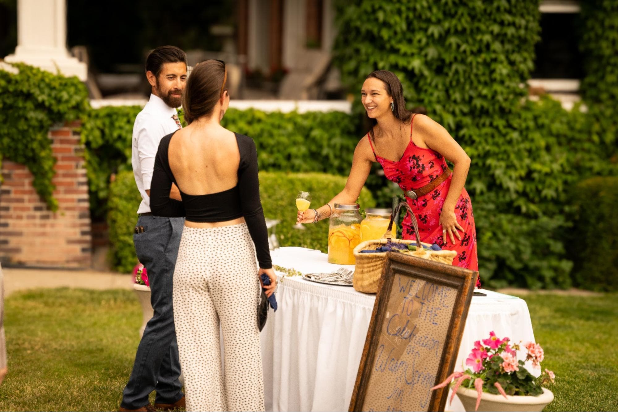 A woman in a red floral dress serves drinks at a garden gathering.