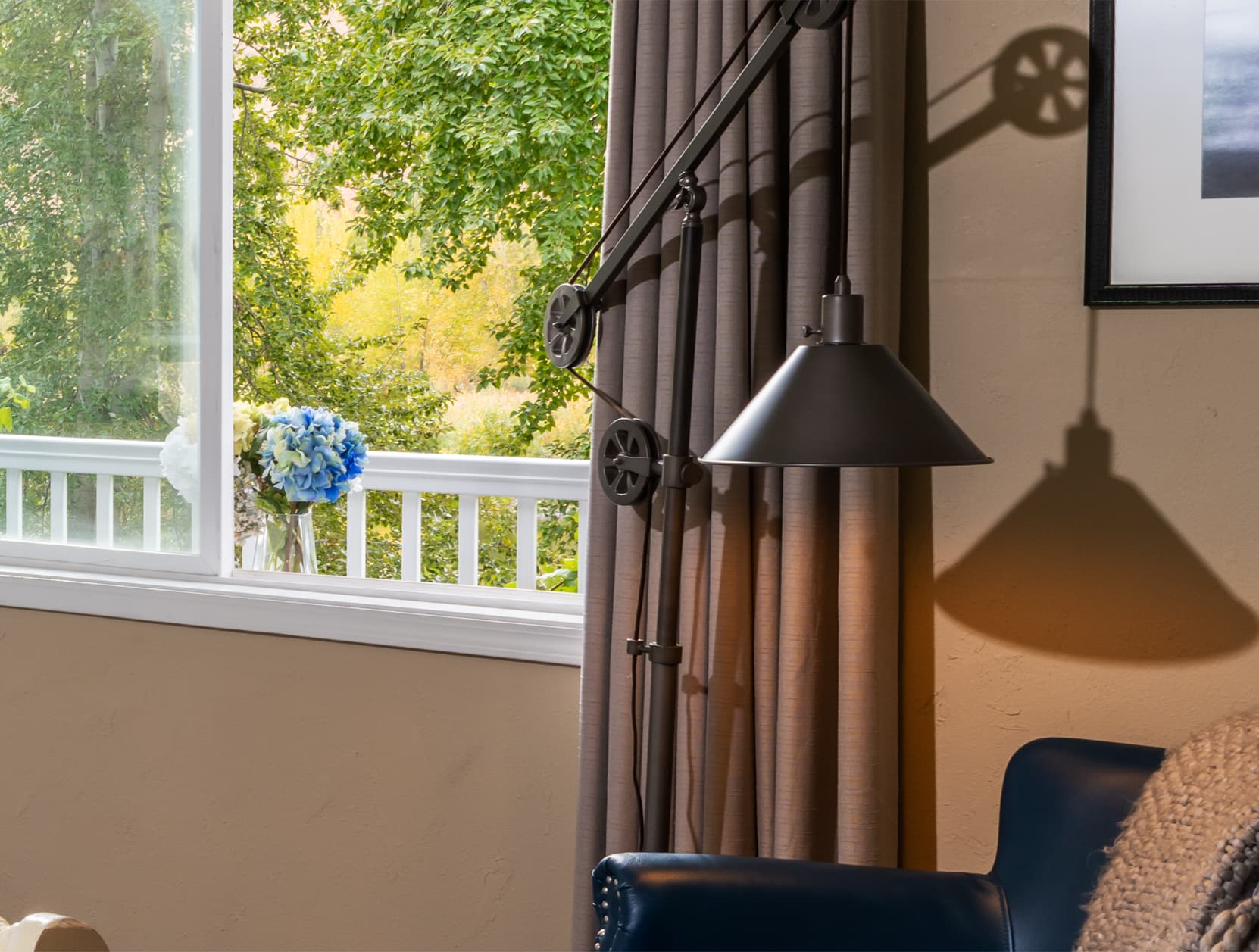 A modern lamp casts a shadow on the wall next to a window with greenery and flowers outside.