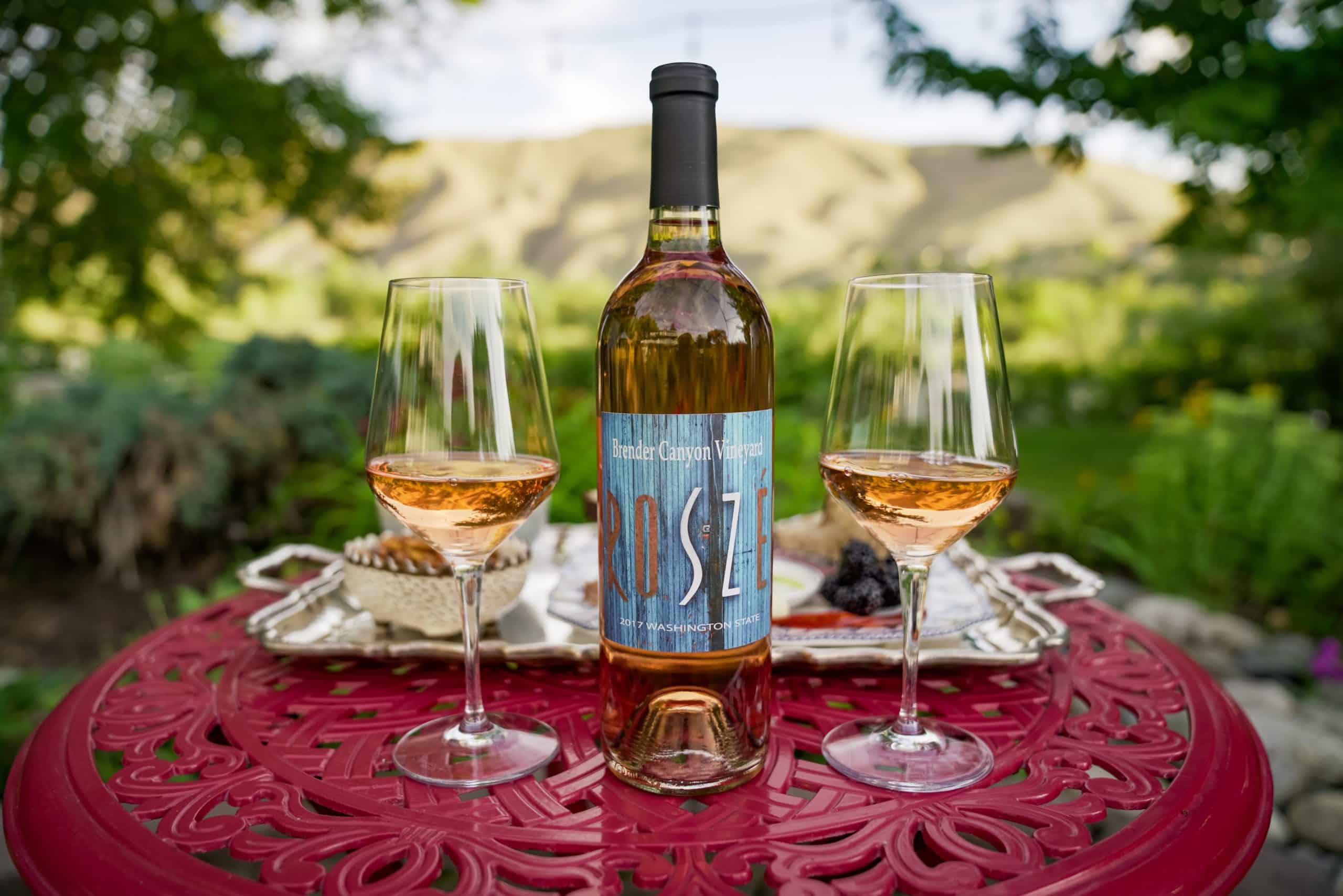 A bottle of rosé wine with two glasses set on a red table in a lush garden.