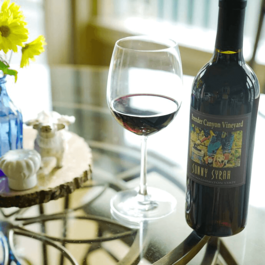 A bottle of Sunny Syrah wine is positioned beside a filled glass on a glass table adorned with flowers and decorative items.