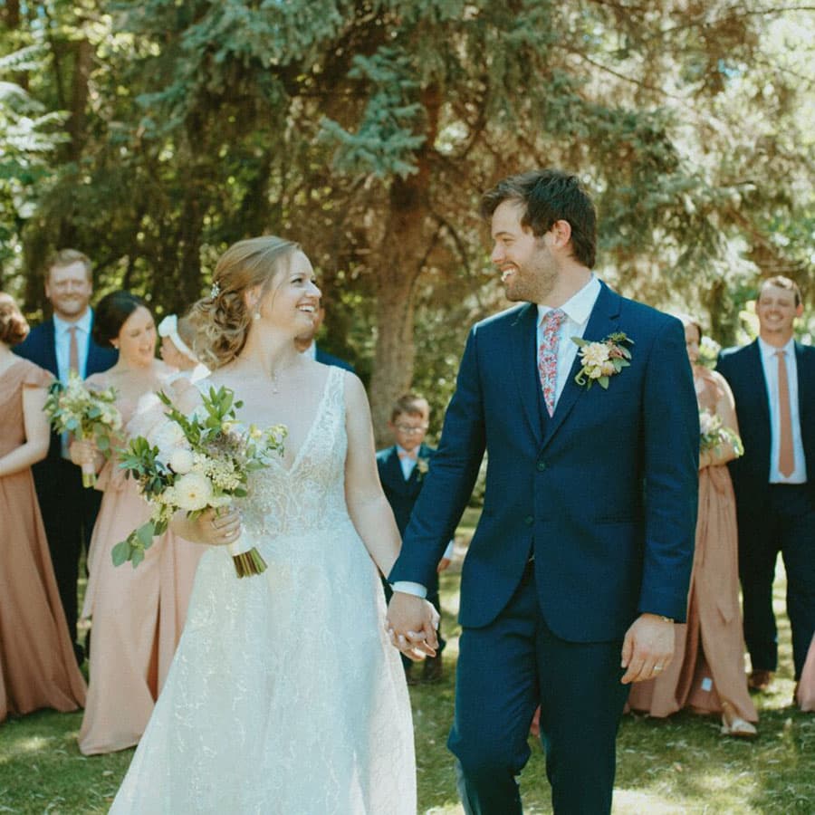 A bride and groom smile at each other while holding hands, surrounded by wedding party members in a forest setting.