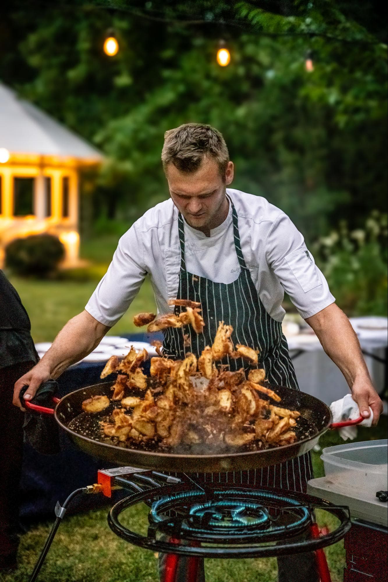 A chef tosses food in a large pan outdoors, illuminated by string lights.