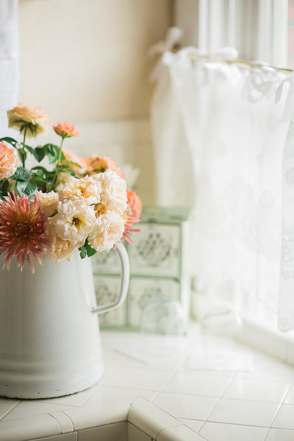 A white pitcher filled with various flowers sits on a countertop beside a window.