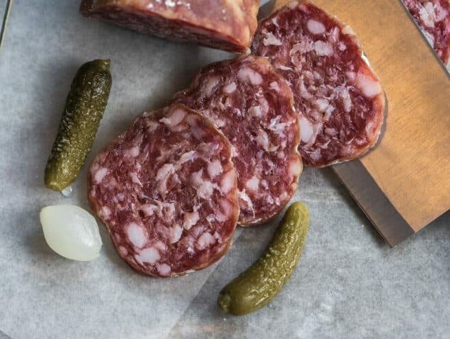 Sliced cured meat accompanied by pickles and shallots on parchment paper.
