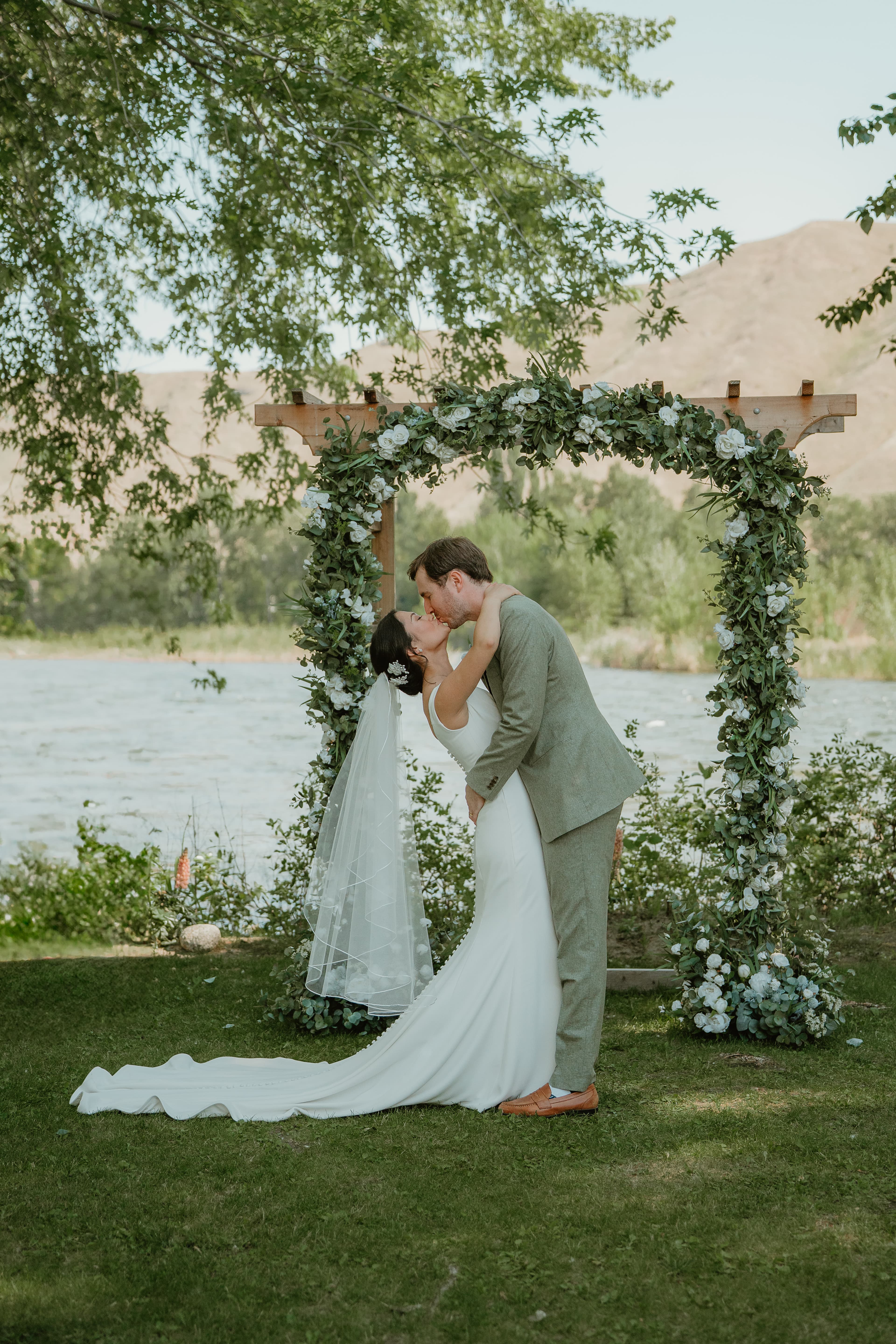 A bride and groom kiss under a floral arch near a river.