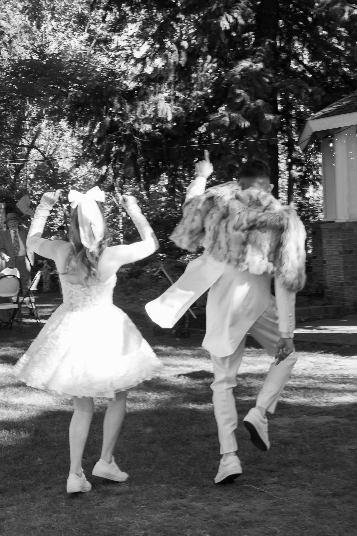 A girl in a white dress and bow dances beside a man in a formal outfit with a fur coat in a sunny outdoor setting.
