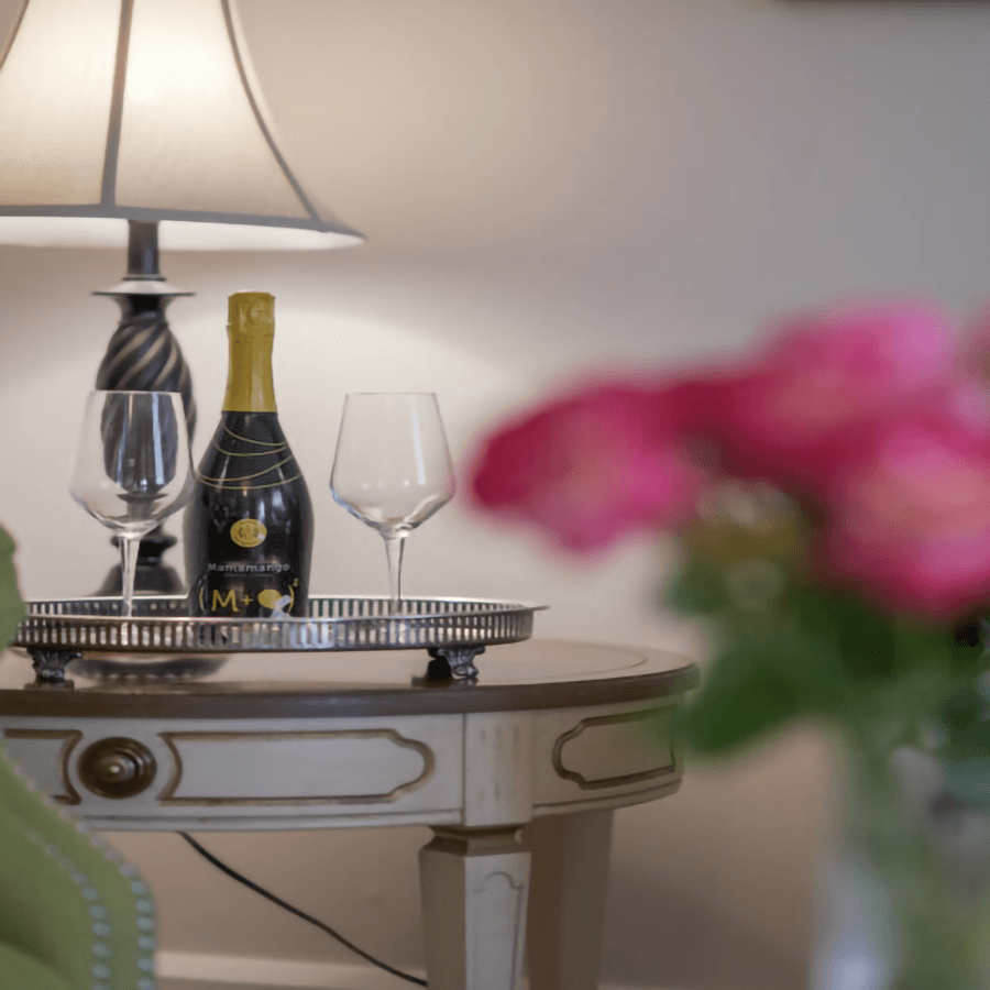 A bottle of sparkling wine and two glasses sit on a tray atop a side table, with a lamp glowing softly nearby.