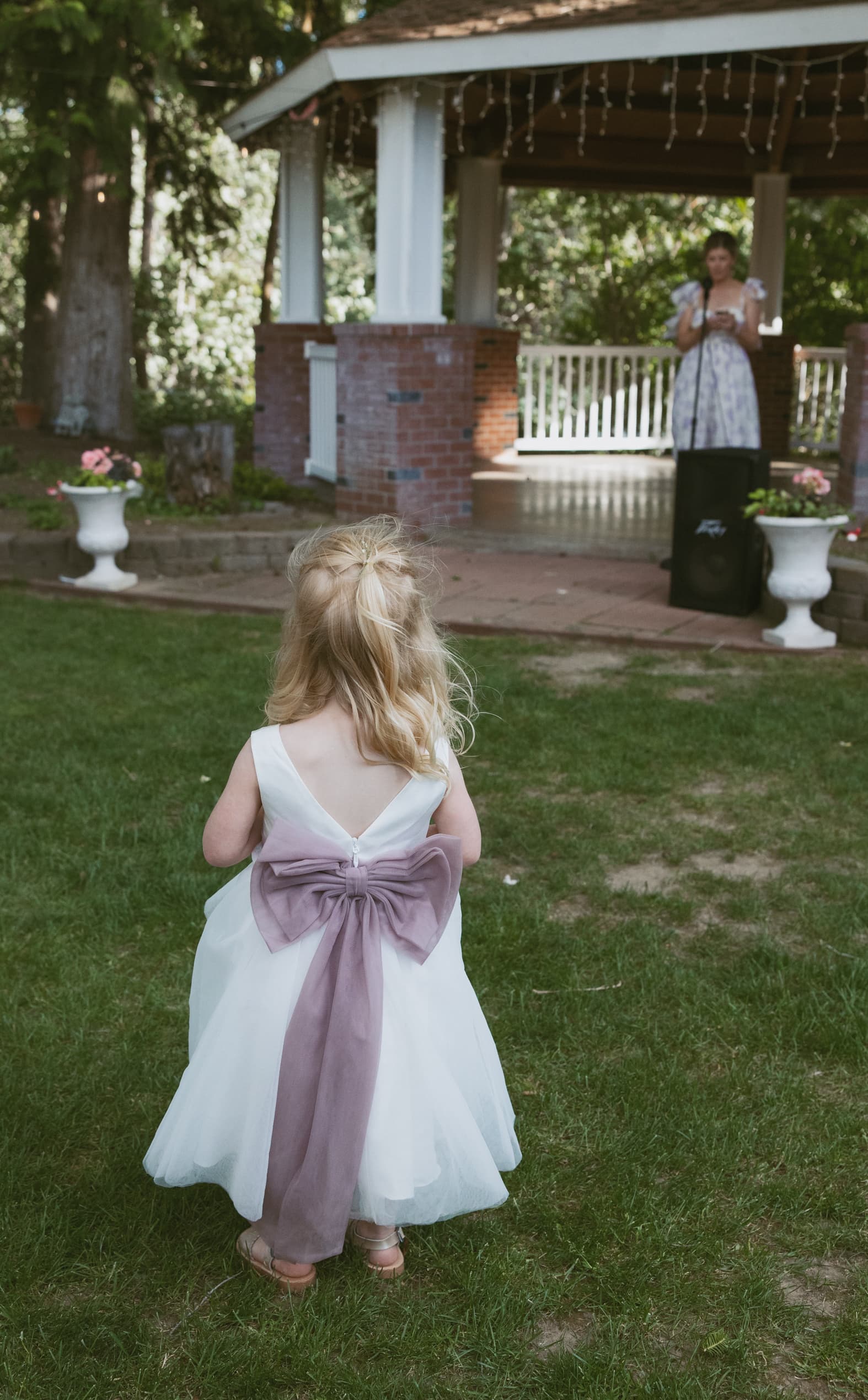 A young girl in a white dress with a purple bow stands with her back to the camera, watching a woman speaking at a gazebo.