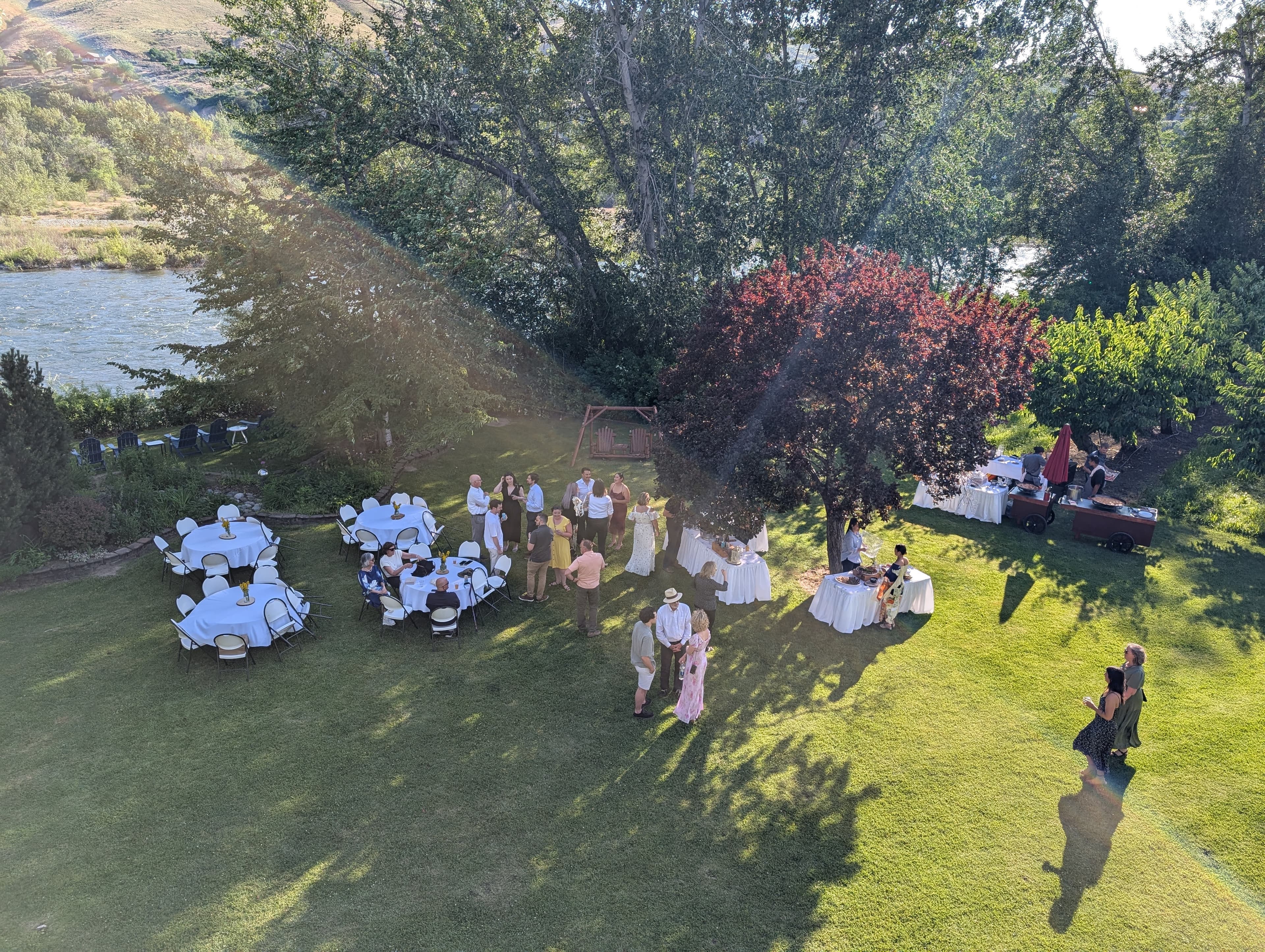 An aerial view of a garden event with tables, guests mingling, and a river in the background.