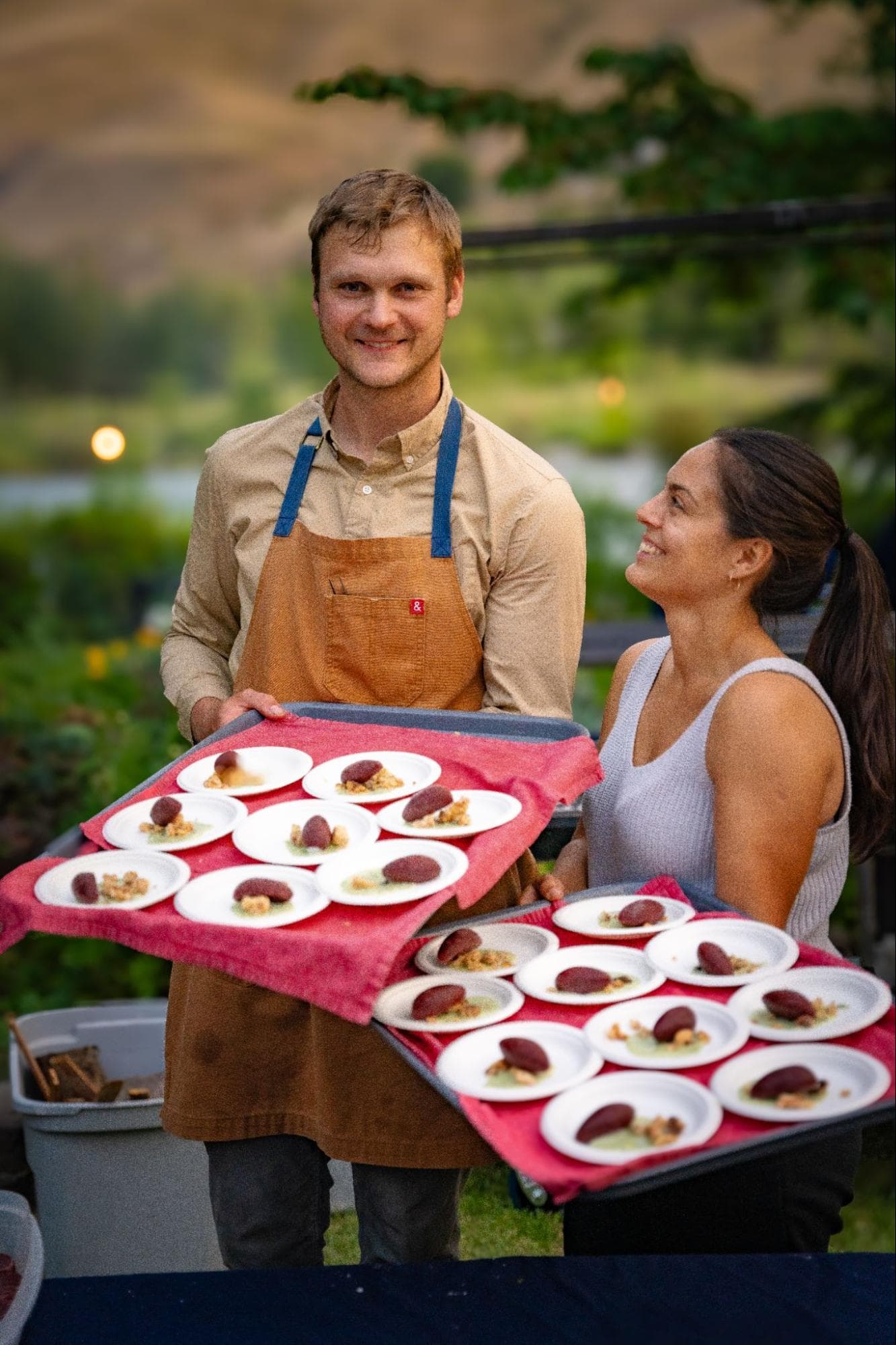 A man and a woman smile while holding trays of beautifully plated desserts outdoors.