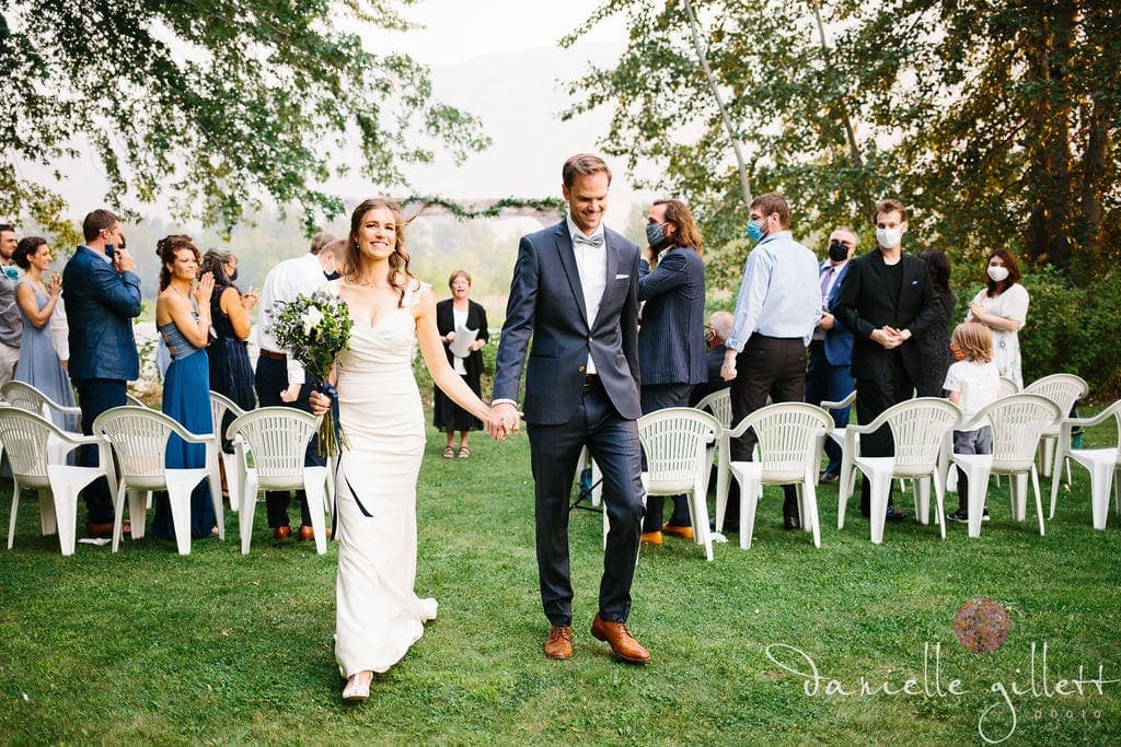 A bride and groom joyfully walk hand in hand down an outdoor aisle surrounded by guests.