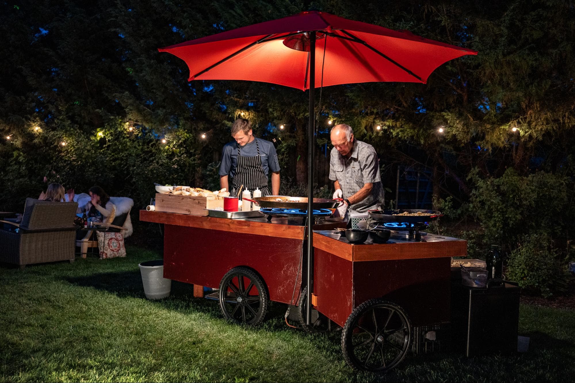 Two chefs cook at a cart under a red umbrella in a softly lit backyard.