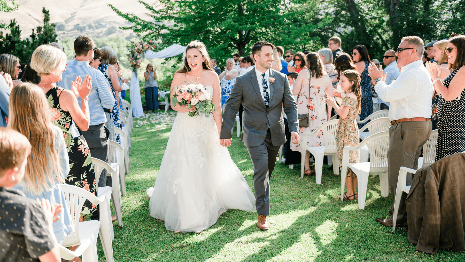 A bride and groom joyfully walk hand-in-hand down the aisle while guests applaud in a lush outdoor setting.