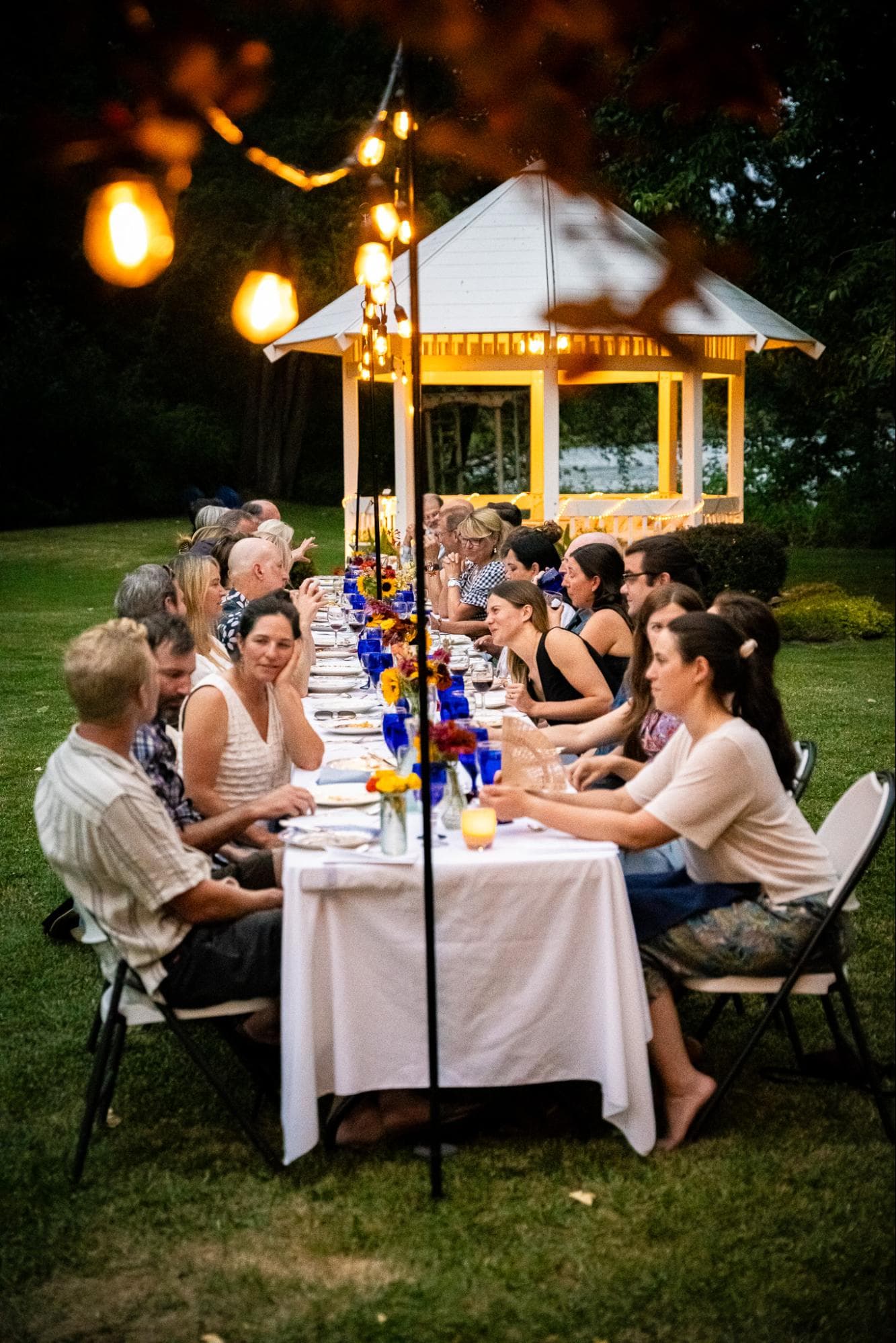 A long outdoor dining table is set up with guests enjoying a meal under warm lights near a gazebo.