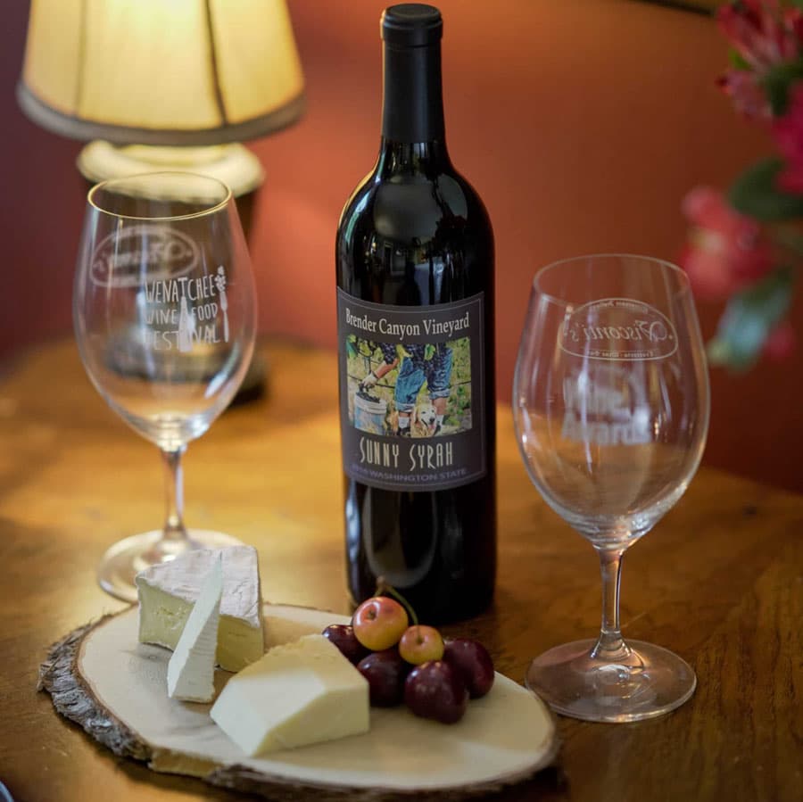 A bottle of Brender Canyon Vineyard Sunny Syrah with two glasses, cheese, and fresh fruit set on a wooden table.