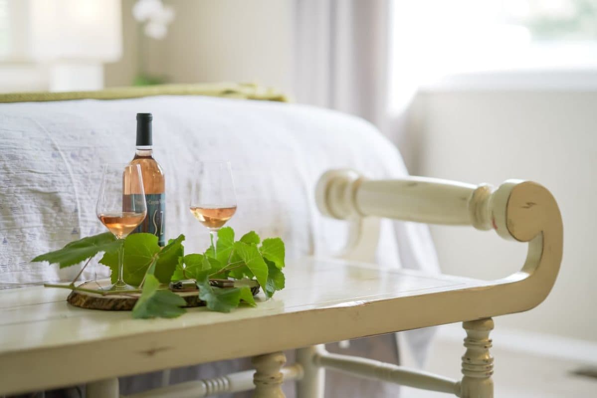 A bottle of rosé and two glasses sit on a wooden table adorned with green leaves, with a softly lit bedroom in the background.